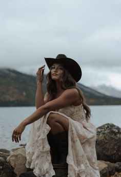 Woman in a cowboy hat and floral dress crouching by a lake with mountains in the background