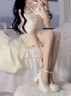 Woman with long black hair in a white dress and high heels sitting on a bed