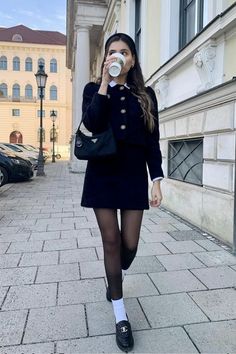 Woman in a black skirt suit and headband walking on a street while drinking coffee