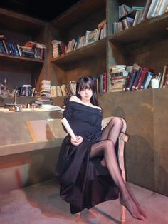 Woman in a black dress and sheer stockings sitting in a room with bookshelves