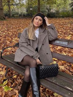 Woman in a plaid blazer and black headband sitting on a park bench with autumn leaves