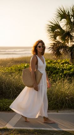 Woman in a white dress and sunglasses walking barefoot on a path near the beach