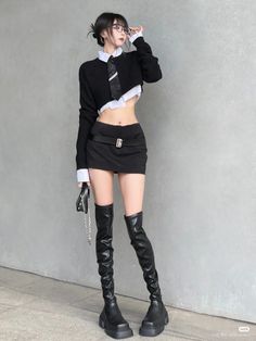 Woman in a black crop top, mini skirt, and thigh high boots against a gray wall