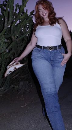 Woman with red hair in a white tank top and blue jeans holding a cowboy hat outdoors at night
