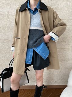 Person in a trench coat, denim shirt, sweater vest, and leather shorts with a tote bag