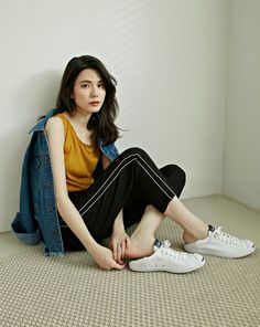 Woman with long dark hair wearing a denim jacket and yellow tank top, sitting indoors