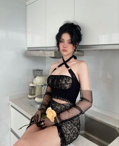 Woman with black hair in a lace outfit and sheer gloves holding a flower in a kitchen