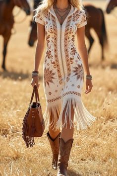 Woman in a fringe dress and cowboy boots walking through dry grass with horses in the background