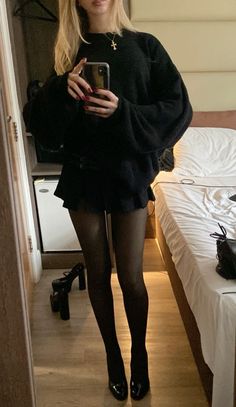 Woman in a black sweater and mini skirt taking a mirror selfie in a room