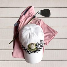 Golf club, white hat with checkered ribbon, and pink shirt on a white wooden surface