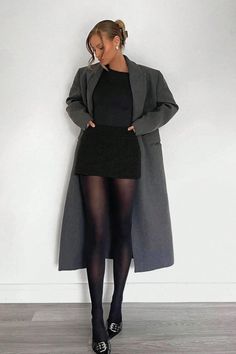 Woman in a long gray coat, black skirt, and black tights posing indoors