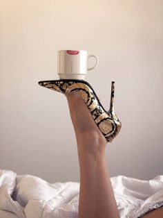 A high heel balancing a coffee cup with a lipstick mark on a leg in bed