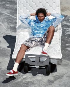 Man with curly hair wearing a blue hoodie and camouflage shorts, sitting on a mattress