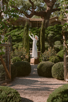 Woman in a white dress and hat standing on a chair in a lush garden