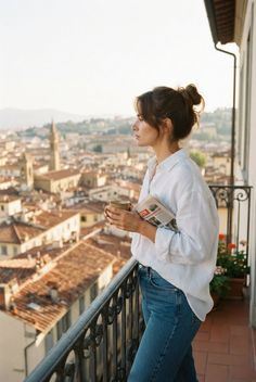 Woman on a balcony overlooking a cityscape, holding a cup and newspaper
