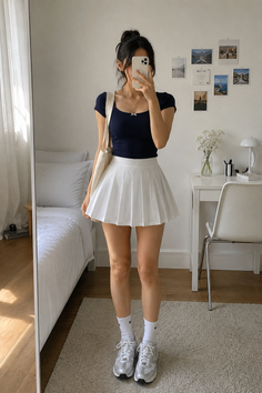 Woman in a navy top and white pleated skirt taking a mirror selfie indoors