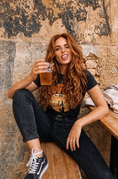 Woman with long curly hair holding a drink, sitting against a rustic wall