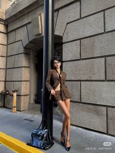 Woman in a brown suit and glasses leaning against a pole on a street