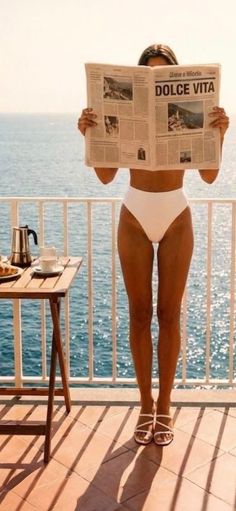 Woman in a white bikini reading a newspaper on a sunny balcony overlooking the ocean
