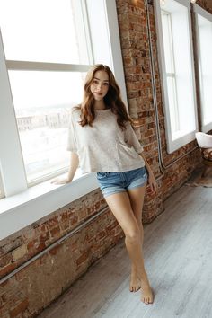 Woman with long hair in denim shorts and a t-shirt standing by a window