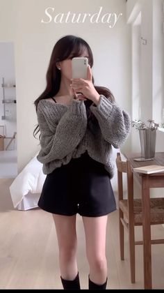 Woman in a gray sweater and black shorts taking a mirror selfie indoors
