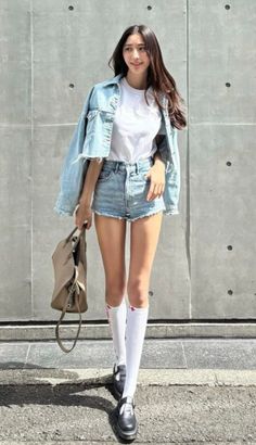 Woman in a denim jacket and shorts with knee-high socks against a concrete wall