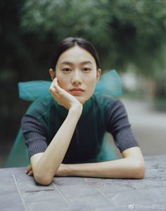 Woman with dark hair wearing a turtleneck shirt and green bow, resting her chin on her hand