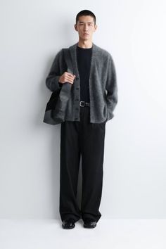 Man in a gray cardigan and black outfit posing against a white wall