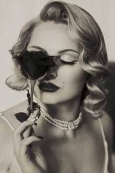 Woman with wavy blonde hair and pearl necklace holding a rose to her face