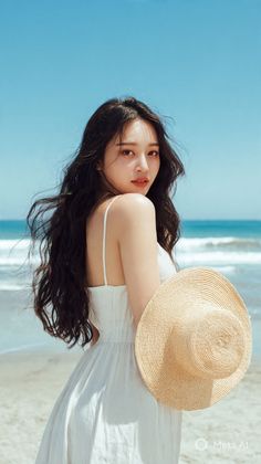 Woman with long dark hair in a white dress holding a straw hat on a beach