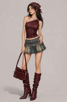 Woman in a one-shoulder top, denim skirt, and knee-high boots holding a handbag