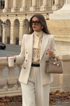 Woman in a cream suit and sunglasses holding a coffee cup and handbag outdoors