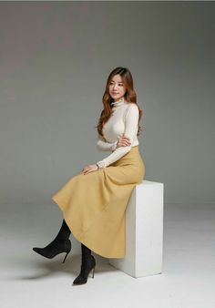 Woman in a cream turtleneck and yellow long skirt sitting on a white block