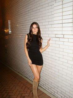 Woman in a black dress and knee-high boots posing against a brick wall at night