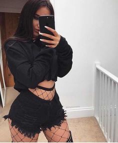 Woman in a black crop top and distressed shorts with fishnet stockings taking a mirror selfie