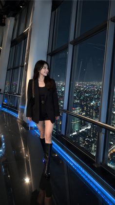 Woman in a black blazer and shorts looking out at a night cityscape from indoors
