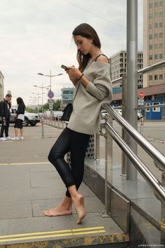Woman in an off-shoulder sweater and black leggings standing barefoot on a city street