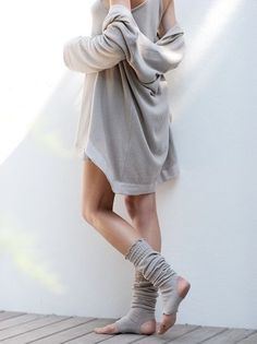 Person in an oversized sweater and leg warmers leaning against a white wall