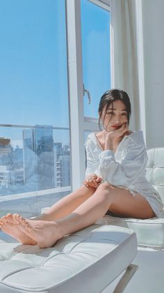 Woman in a white shirt dress sitting on a white couch by a window overlooking a cityscape