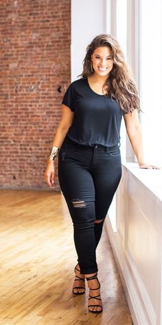 Woman with long wavy hair in a black t-shirt and ripped jeans, smiling indoors