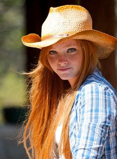 Woman with long red hair and freckles wearing a straw hat and plaid shirt