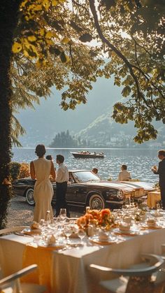 People gathered by a lake with a vintage car and a set table at golden hour