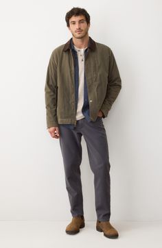 Man in an olive jacket, denim shirt, and dark pants standing against a white wall