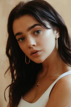 Woman with long dark hair wearing a white tank top and gold earrings