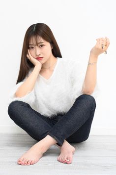 Woman with long hair wearing a white fluffy top and dark jeans, sitting barefoot indoors