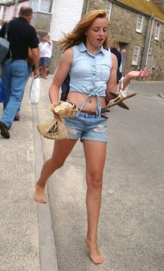 Young woman in denim shorts and a tied crop top walking barefoot on a street