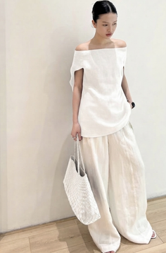 Woman in a white off-shoulder top and wide-leg pants holding a woven bag