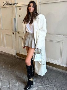 Woman in a white coat, pleated skirt, and black boots holding a mini bag