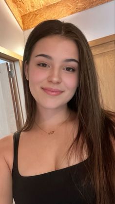 Young woman with long brown hair wearing a black top and gold necklace indoors