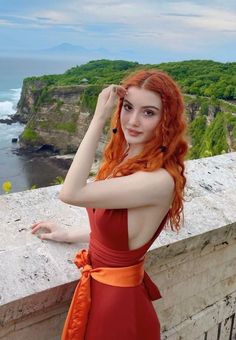 Woman with red hair in a red dress posing on a cliff overlooking the ocean